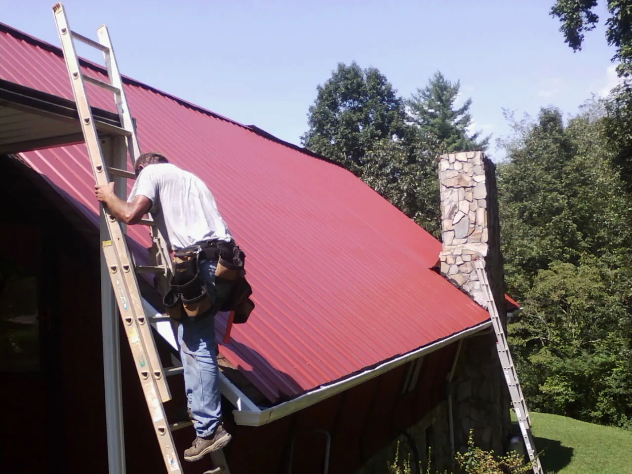 Roofer on a ladder beside a red metal roof and stone chimney — Soffit & Fascia Repair by Roofing Brothers in Agness
